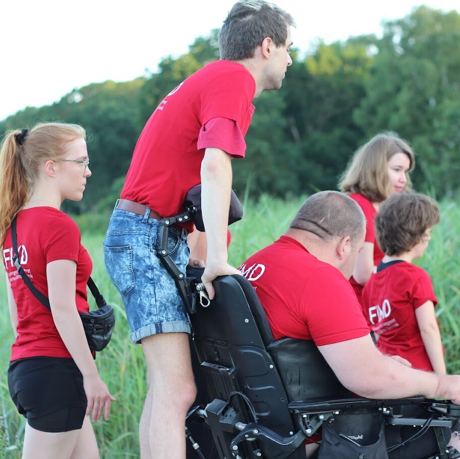 En gruppe deltagere er udendørs i skoven. En af deltagerene står bagpå en anden deltagers kørestol.
